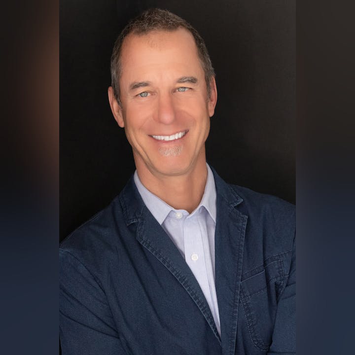 A person smiling in a denim blazer and a light shirt, set against a dark background.