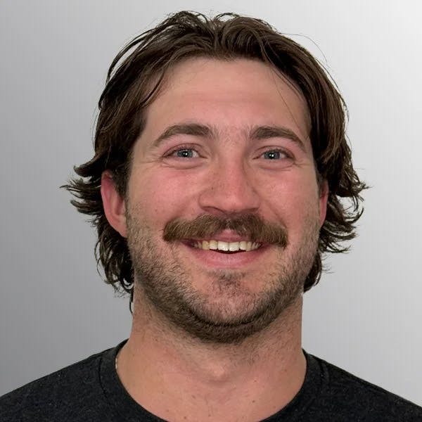 A person with long hair and a mustache smiling against a plain background.