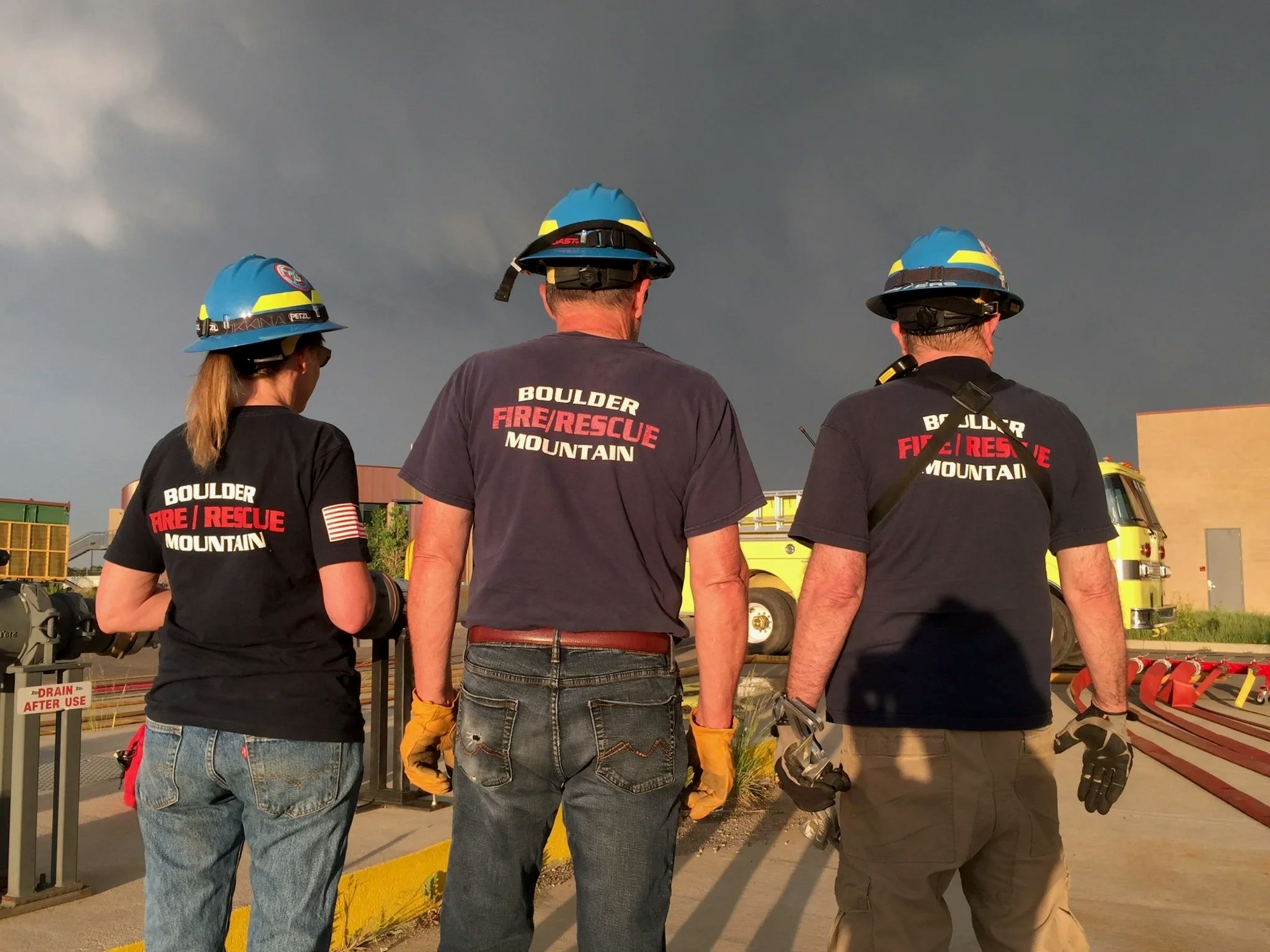 Three firefighters wearing helmets and "Boulder Fire Rescue Mountain" shirts, standing in front of a fire truck.