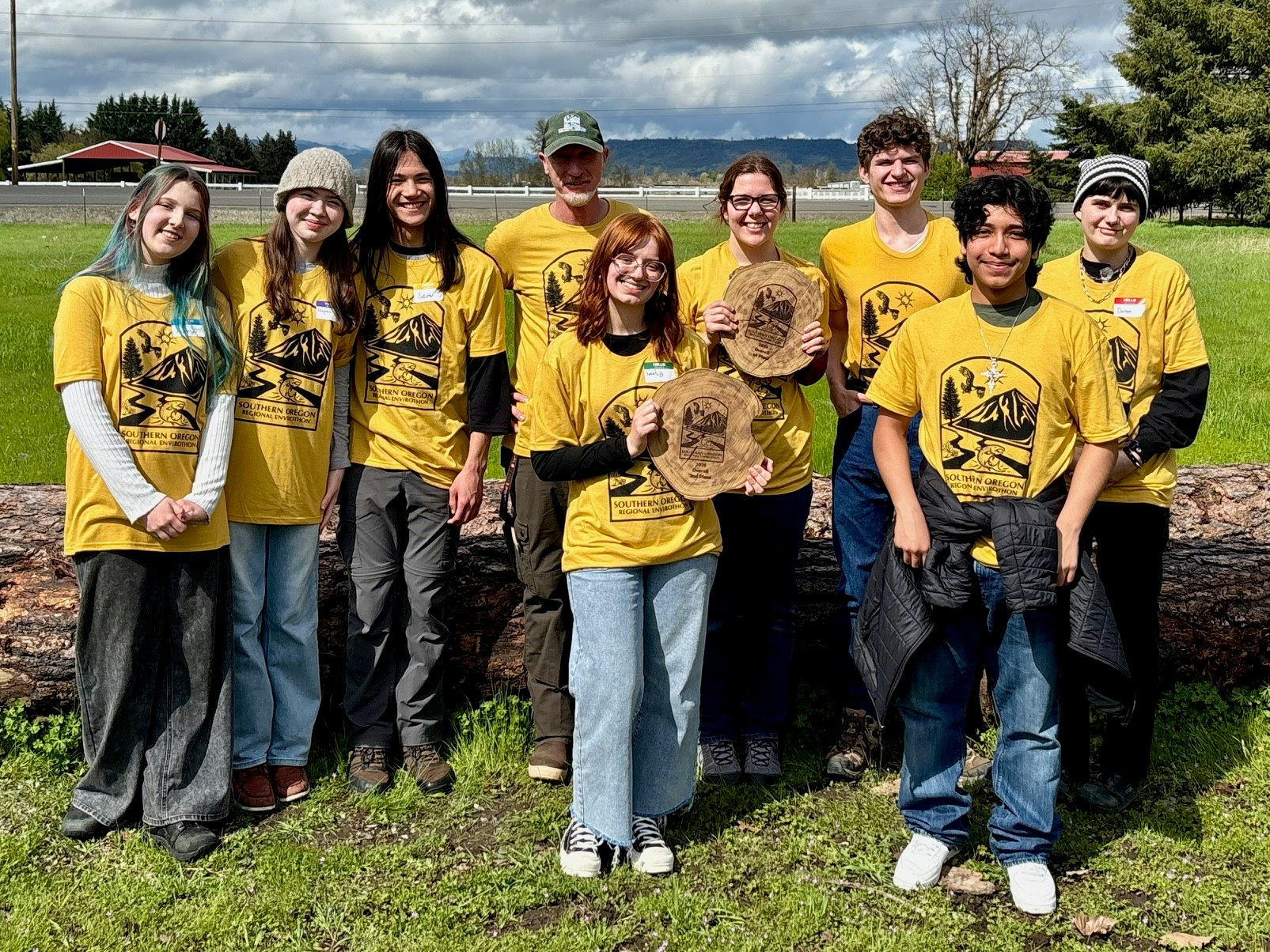 A group of people in yellow Southern Oregon shirts, outdoors, holding plaques, with trees and a building in the background.