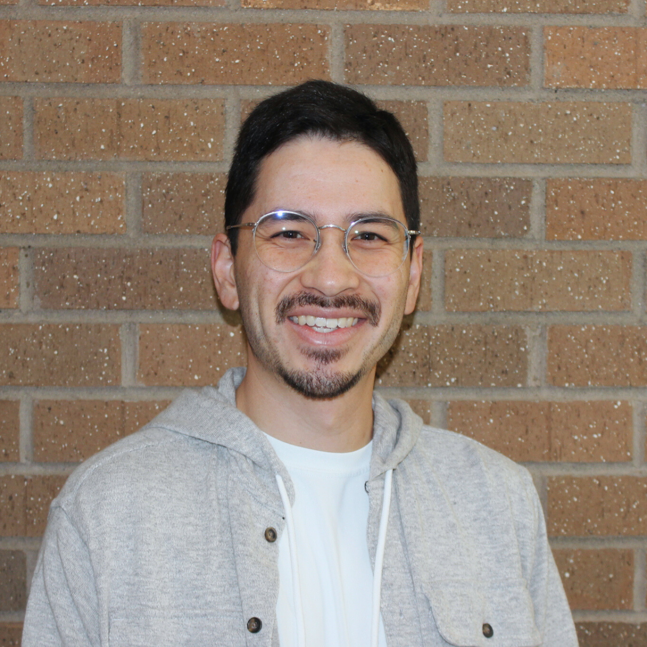A smiling person wearing glasses and a gray hoodie stands in front of a brick wall.