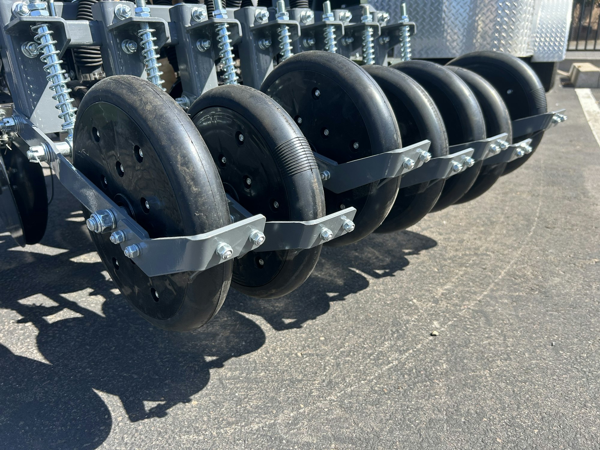 Six black rubber wheels mounted on a metal frame, part of industrial or agricultural machinery, on a paved surface.