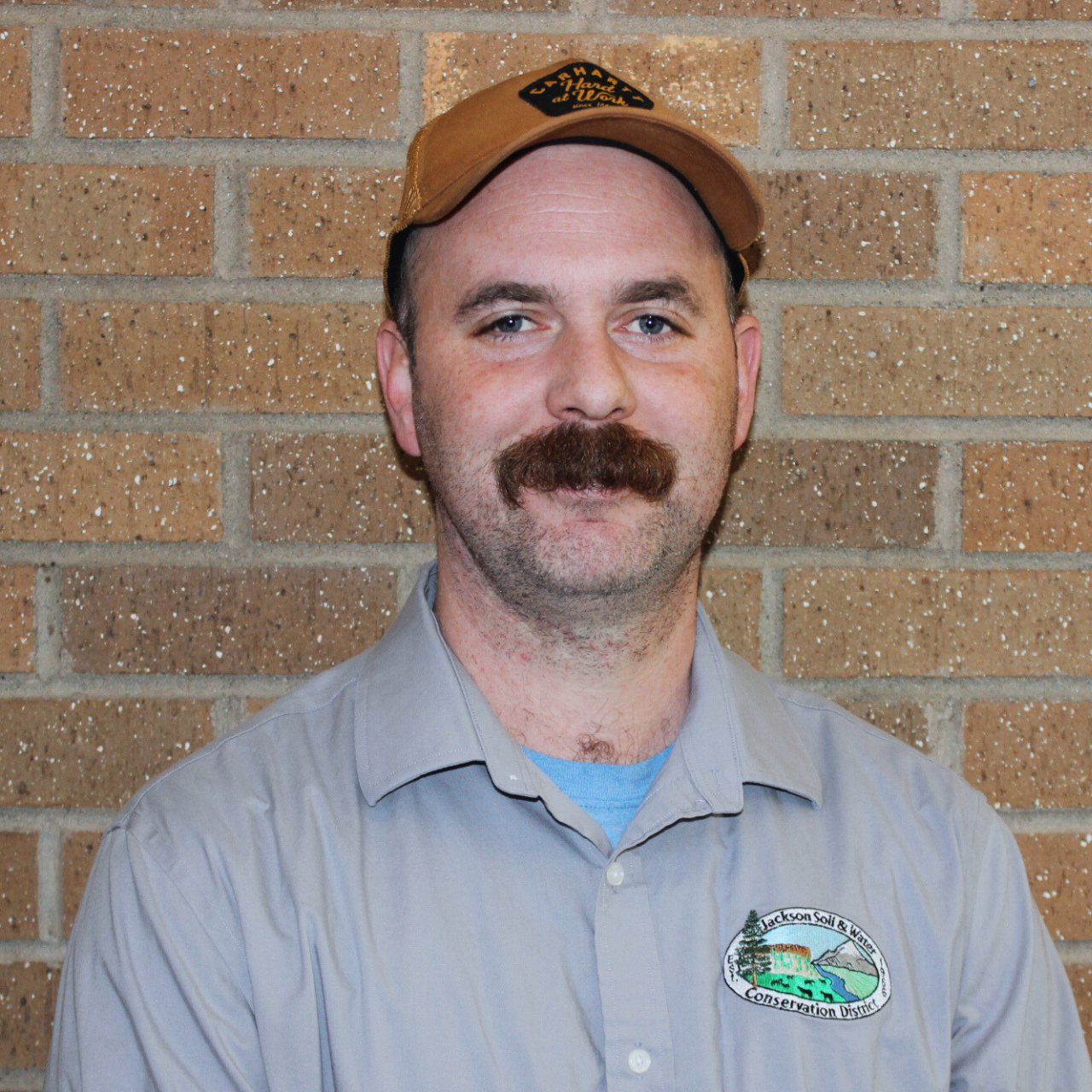 Man with a mustache wearing a cap and a shirt with a patch, standing in front of a brick wall.