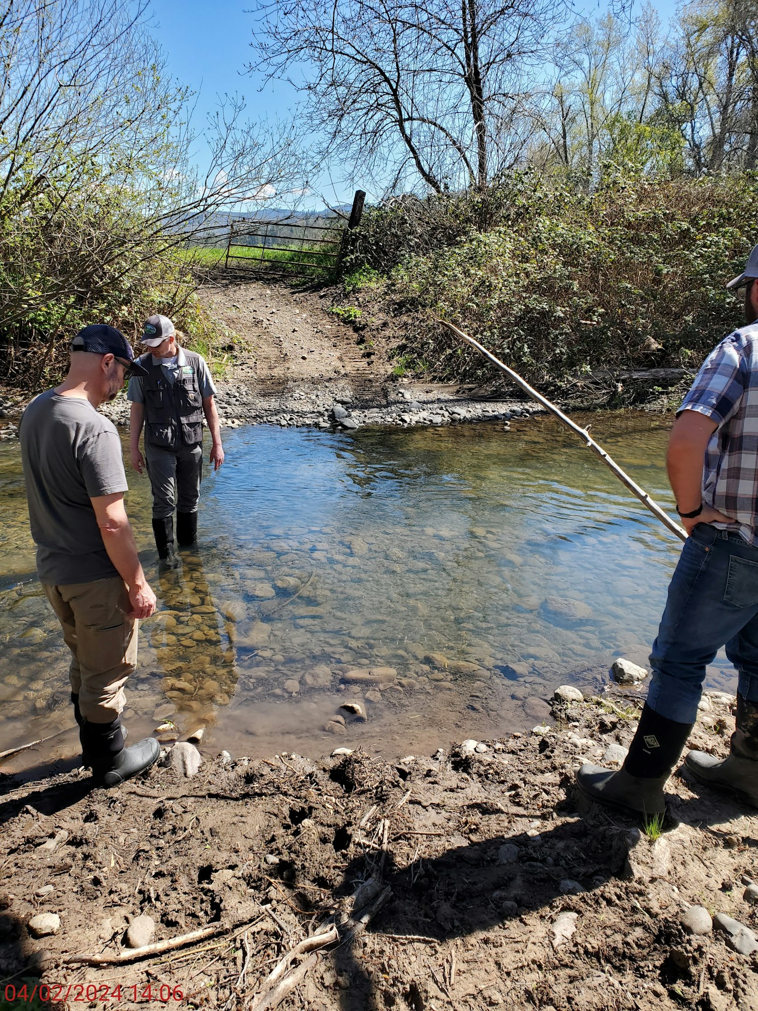 Three people stand by a shallow creek, wearing boots and hats, surrounded by trees and bushes under a clear blue sky.