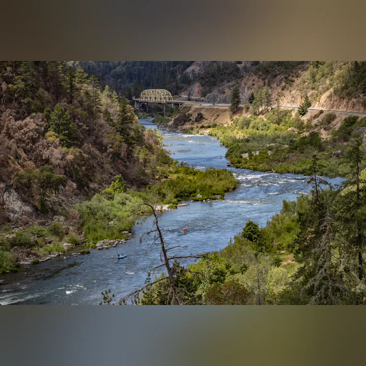 A scenic river flows through a green valley with trees and a bridge, while rafts navigate the water under a blue sky.