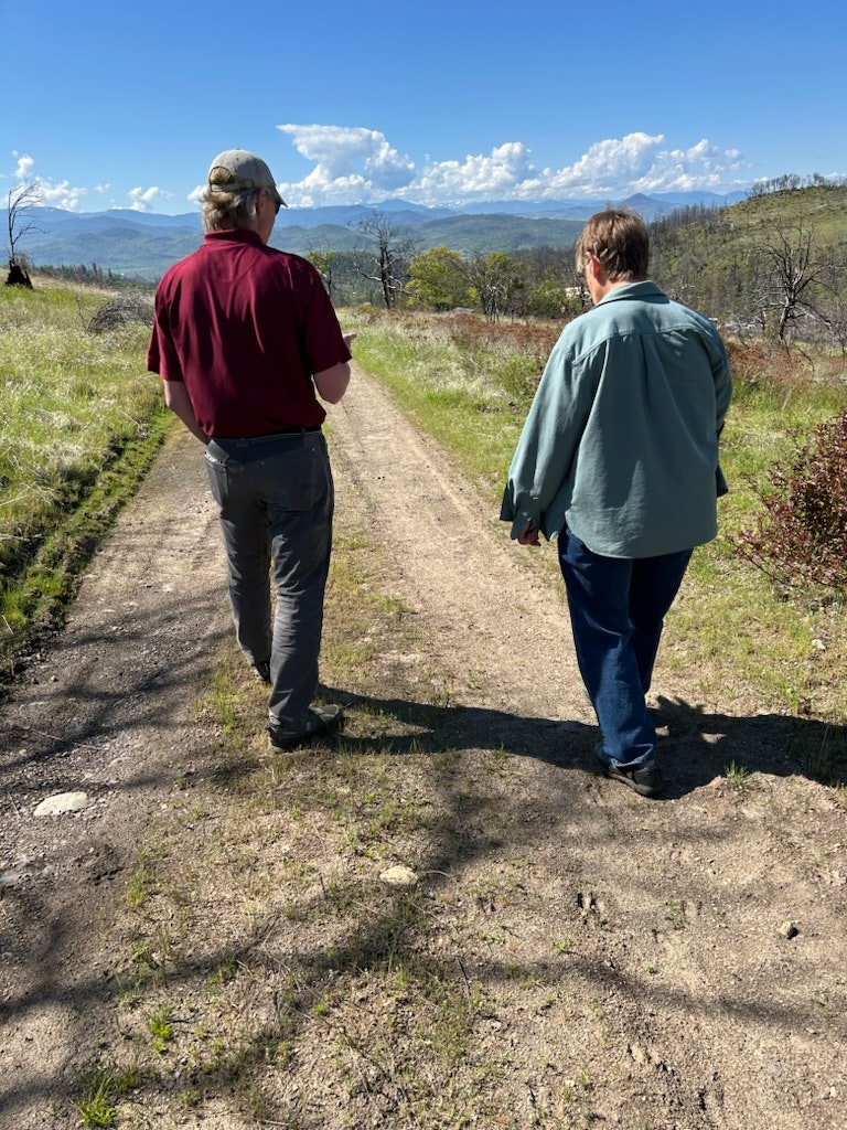 Two people walking on a dirt path in a scenic landscape with mountains and blue skies, surrounded by greenery.