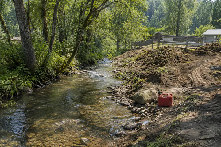 A serene creek surrounded by greenery, with a red gas can and a dirt path nearby. Peaceful nature scene.