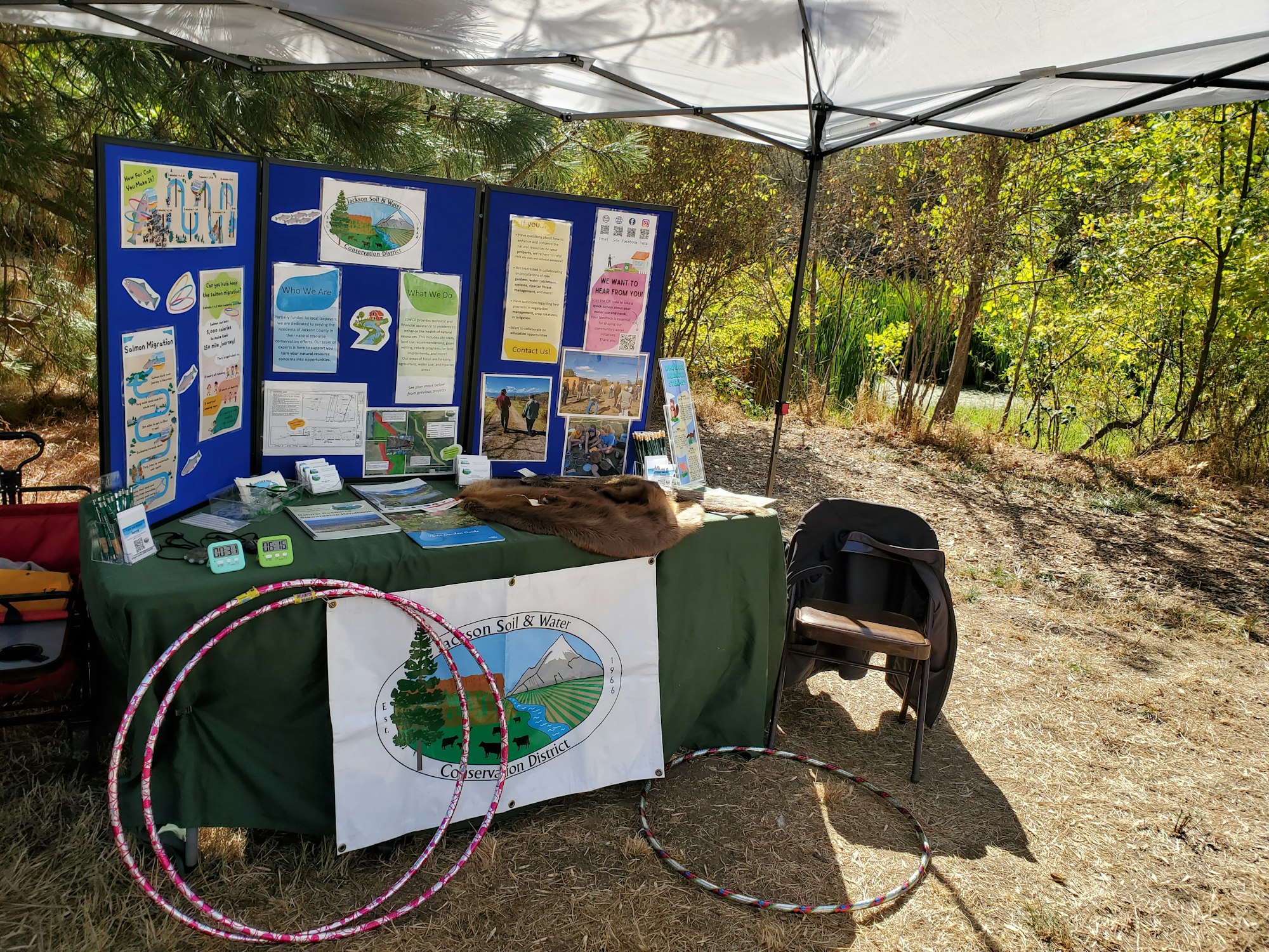 Outdoor information booth with educational displays, papers, timers, hula hoops, and a conservation district banner under a canopy.