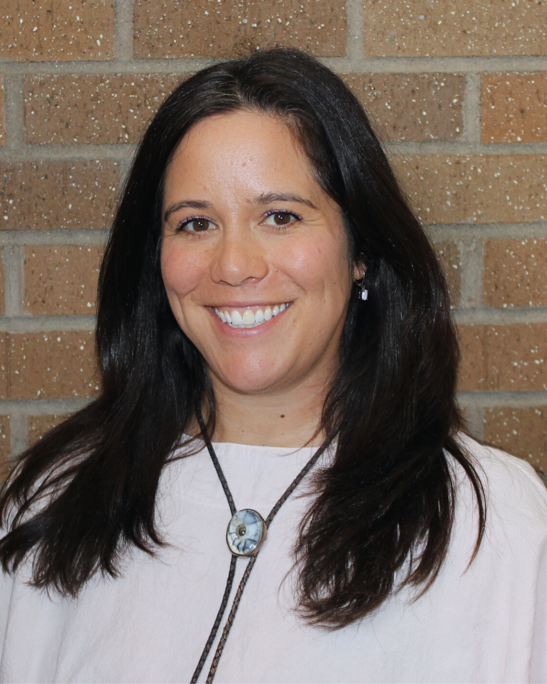 A person with dark hair smiling in front of a brick wall, wearing a light-colored top and a necklace.