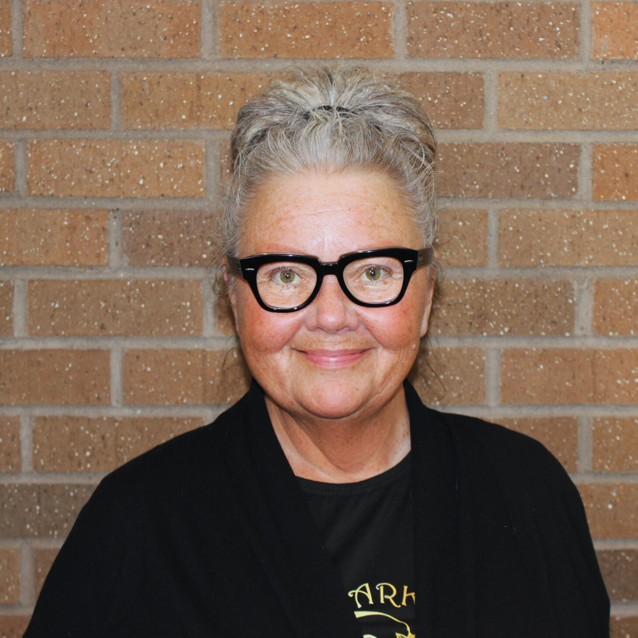 A person with glasses and gray hair, smiling against a brick wall background.