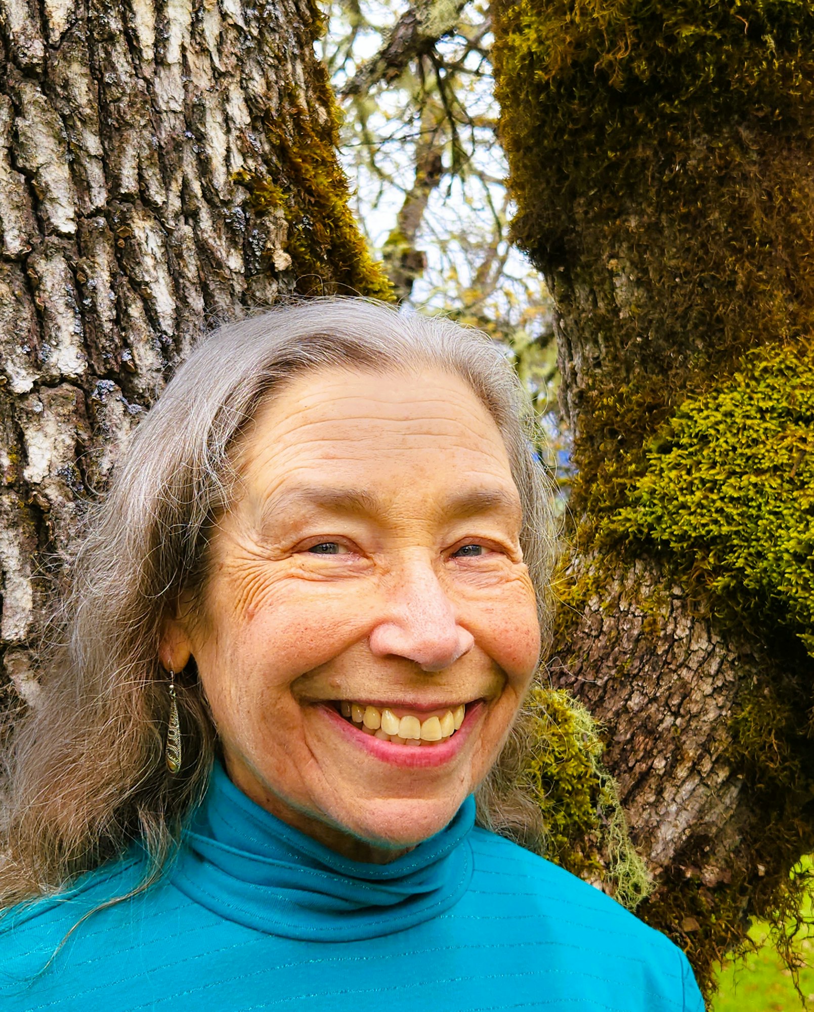 Elderly woman smiling, standing by mossy trees.