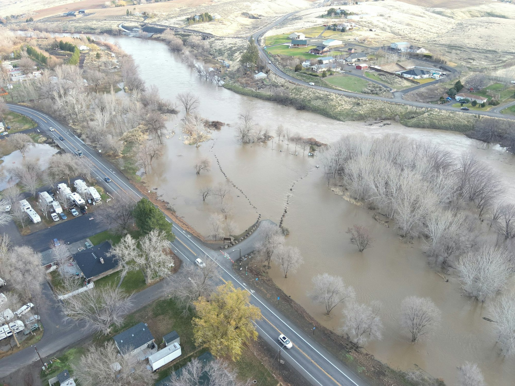 Aerial view of a flooded area with a river, roads, scattered houses, and trees submerged in water, indicating recent heavy rainfall.