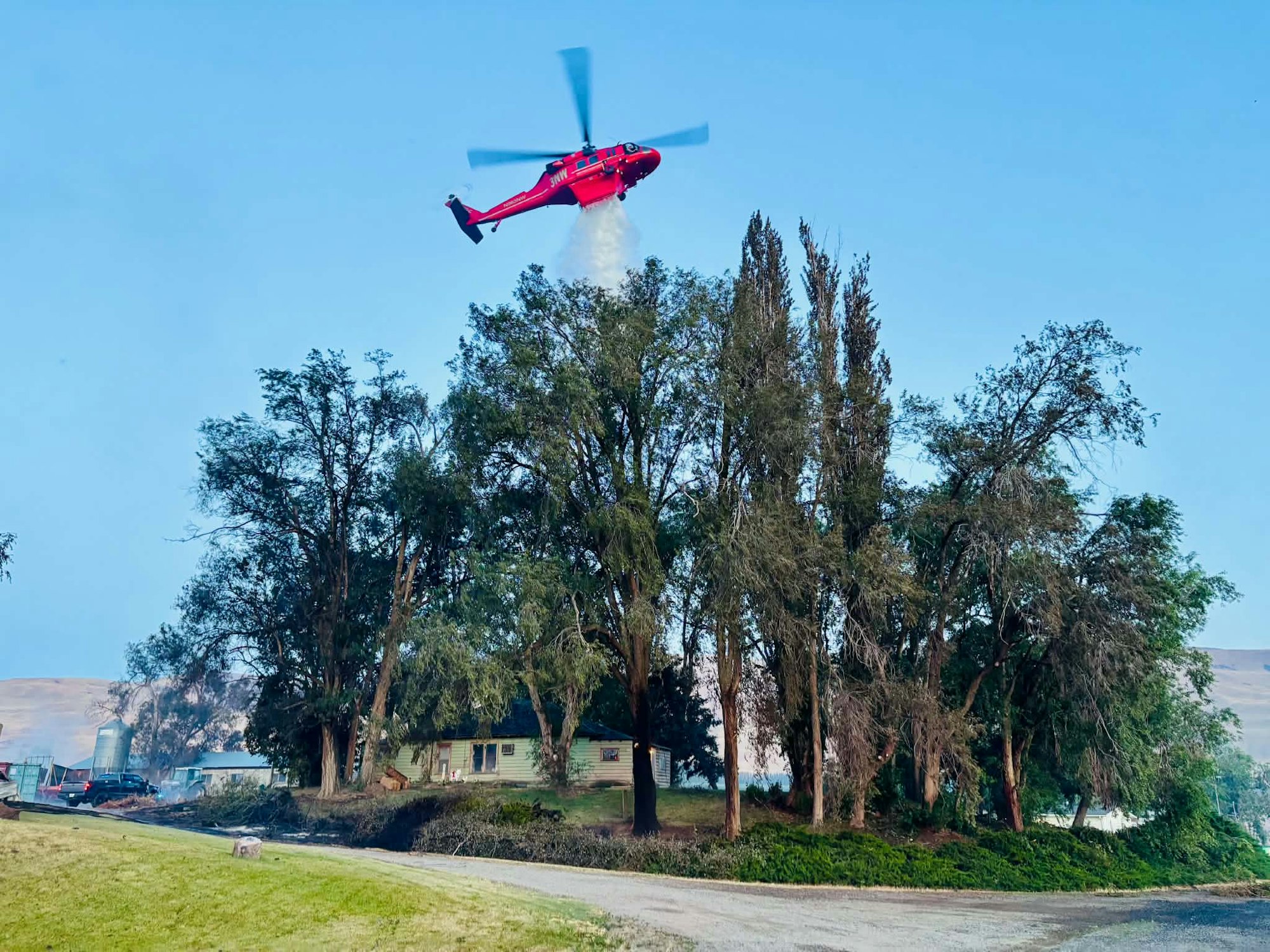 A red helicopter hovers above trees and a house, possibly battling a nearby fire, with smoke visible in the background.