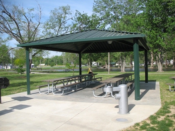 A green-roofed pavilion in a park with picnic tables, a water fountain, and a grill nearby, surrounded by trees and greenery.