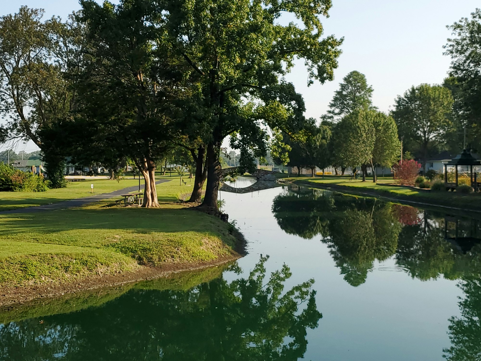 A serene park scene featuring a calm pond, lush trees, and reflected greenery under clear skies.
