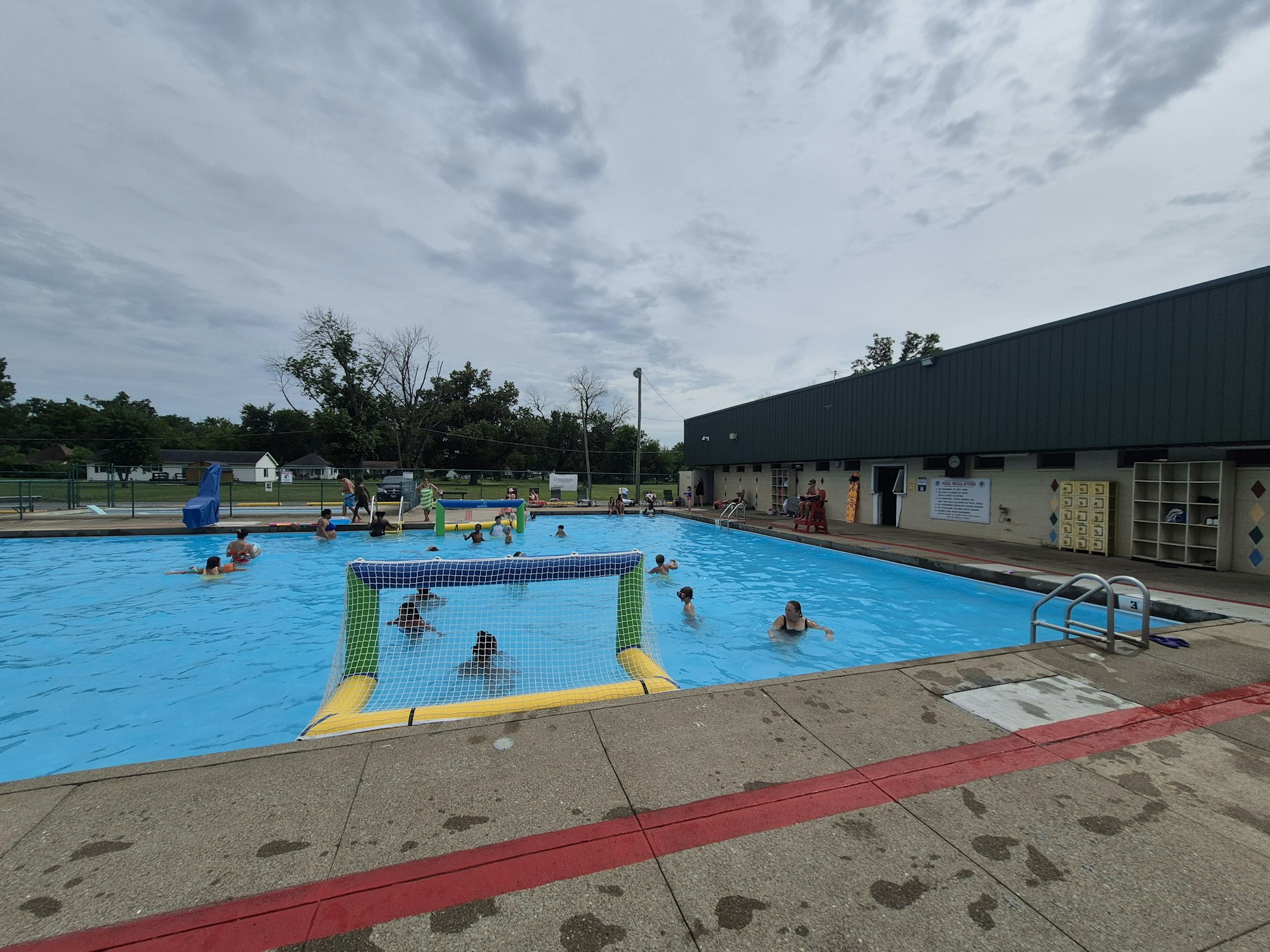 A community pool scene with people swimming, playing, and lounging, surrounded by cloudy skies and green trees.