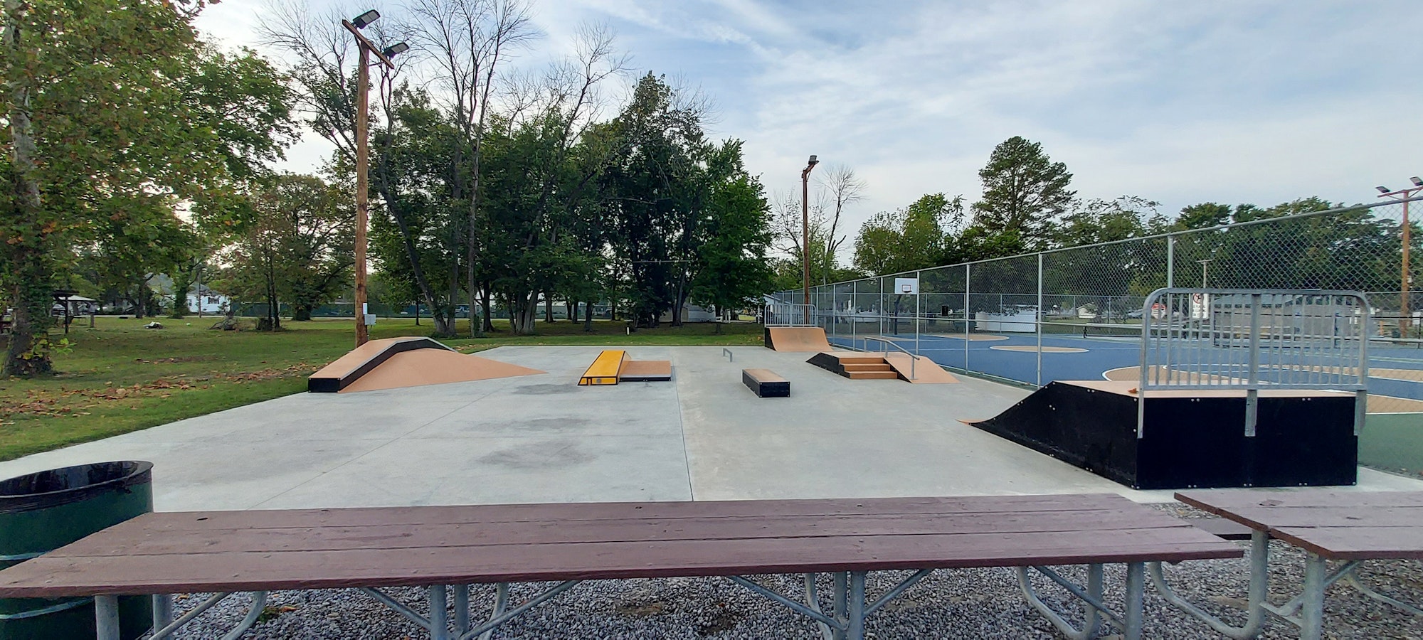 A skatepark featuring ramps and rails, surrounded by greenery and a basketball court nearby. Ideal for skating enthusiasts.