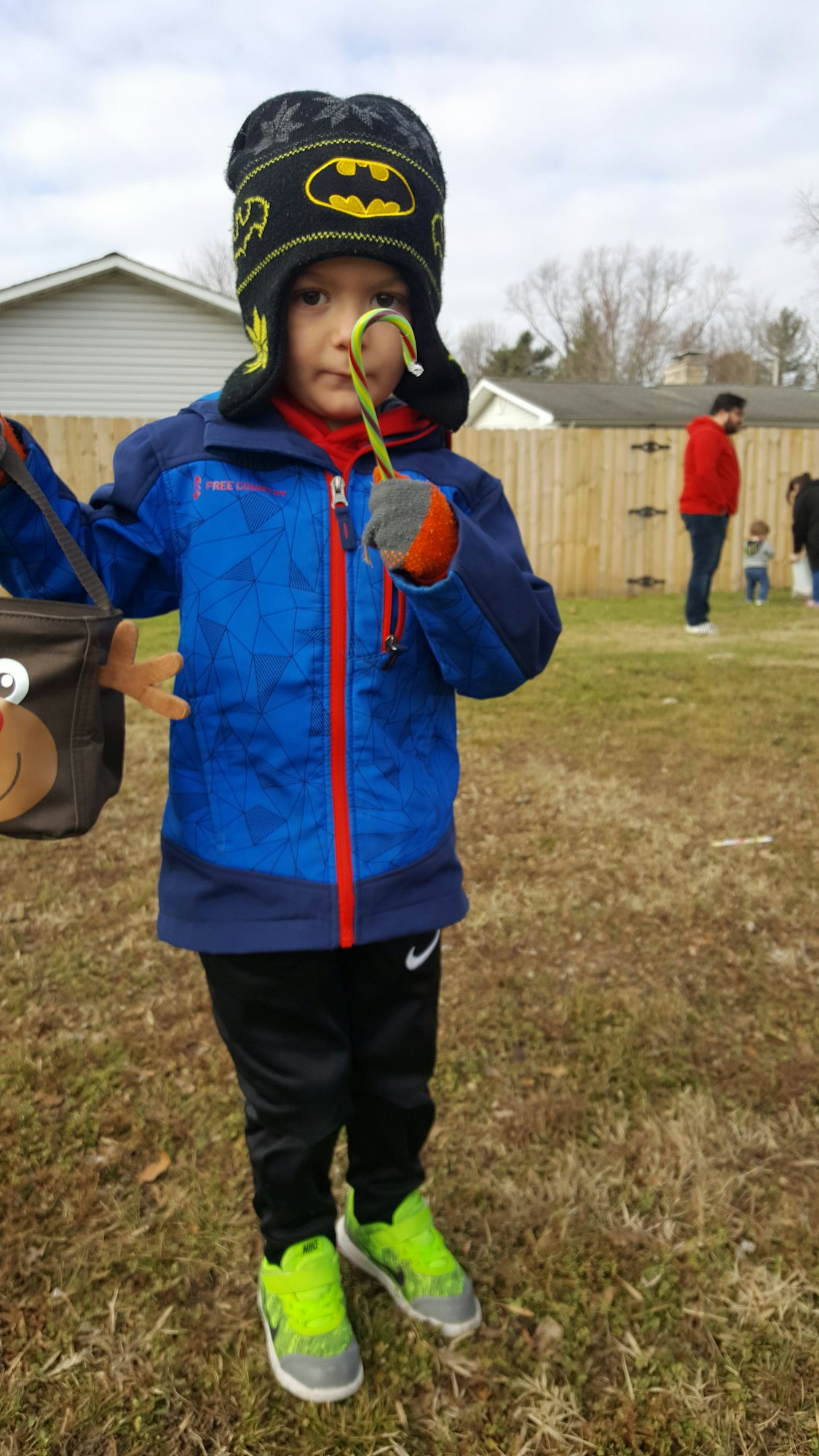 A child in a Batman hat and colorful jacket holds a candy cane, standing on grass with adults in the background.