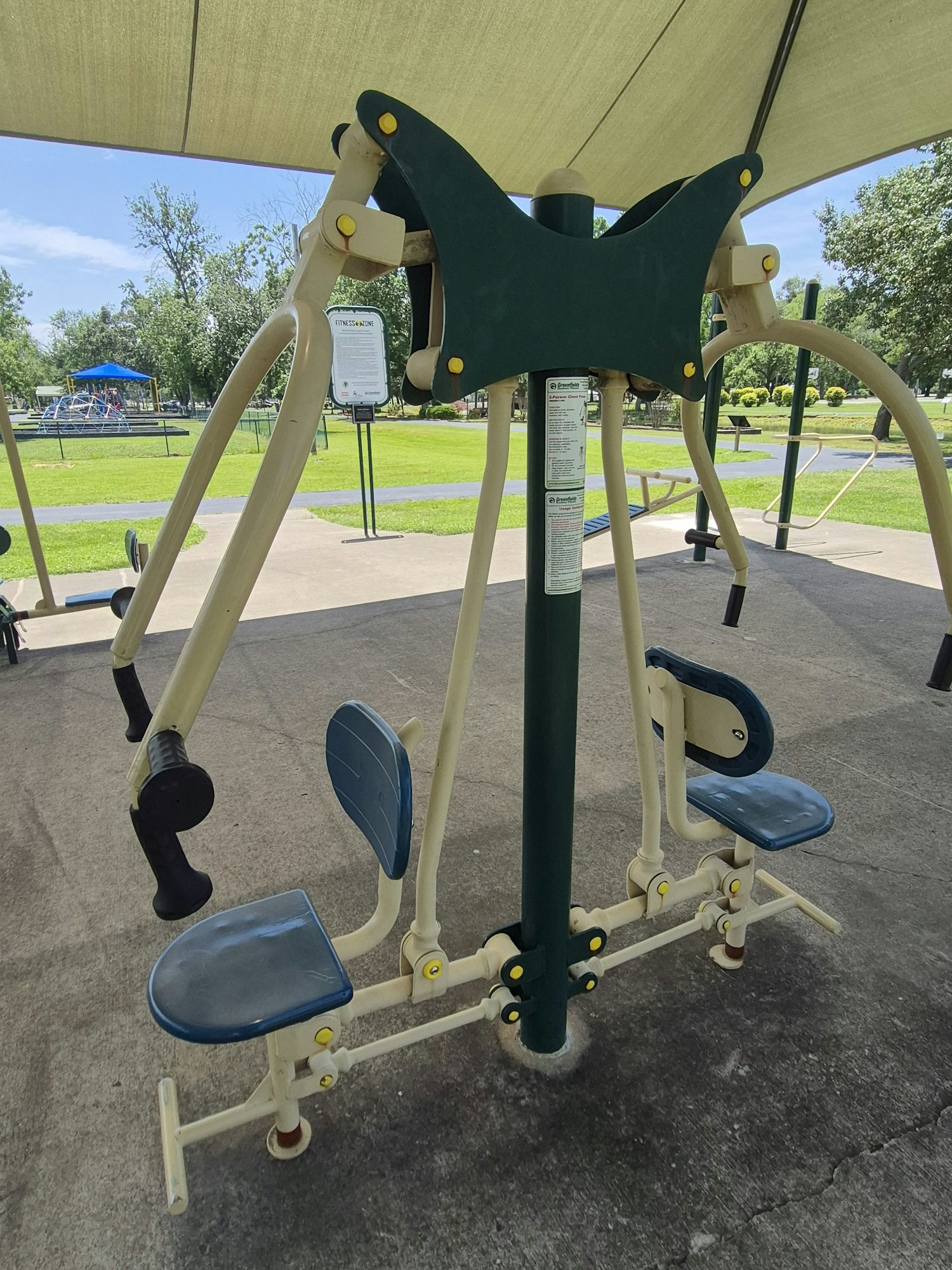 The image shows outdoor gym equipment, likely for exercise and fitness, under a shade structure in a park setting.
