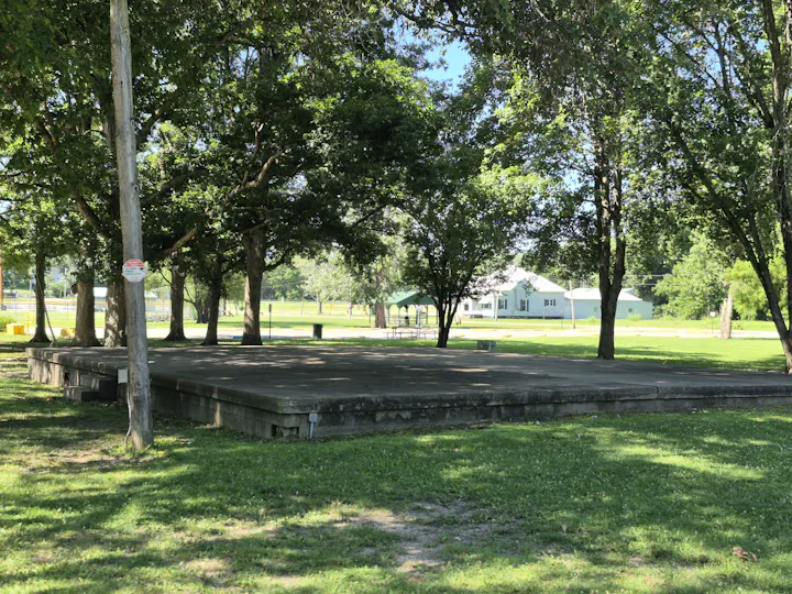A grassy park area with a large concrete stage and trees, featuring a nearby building in the background.