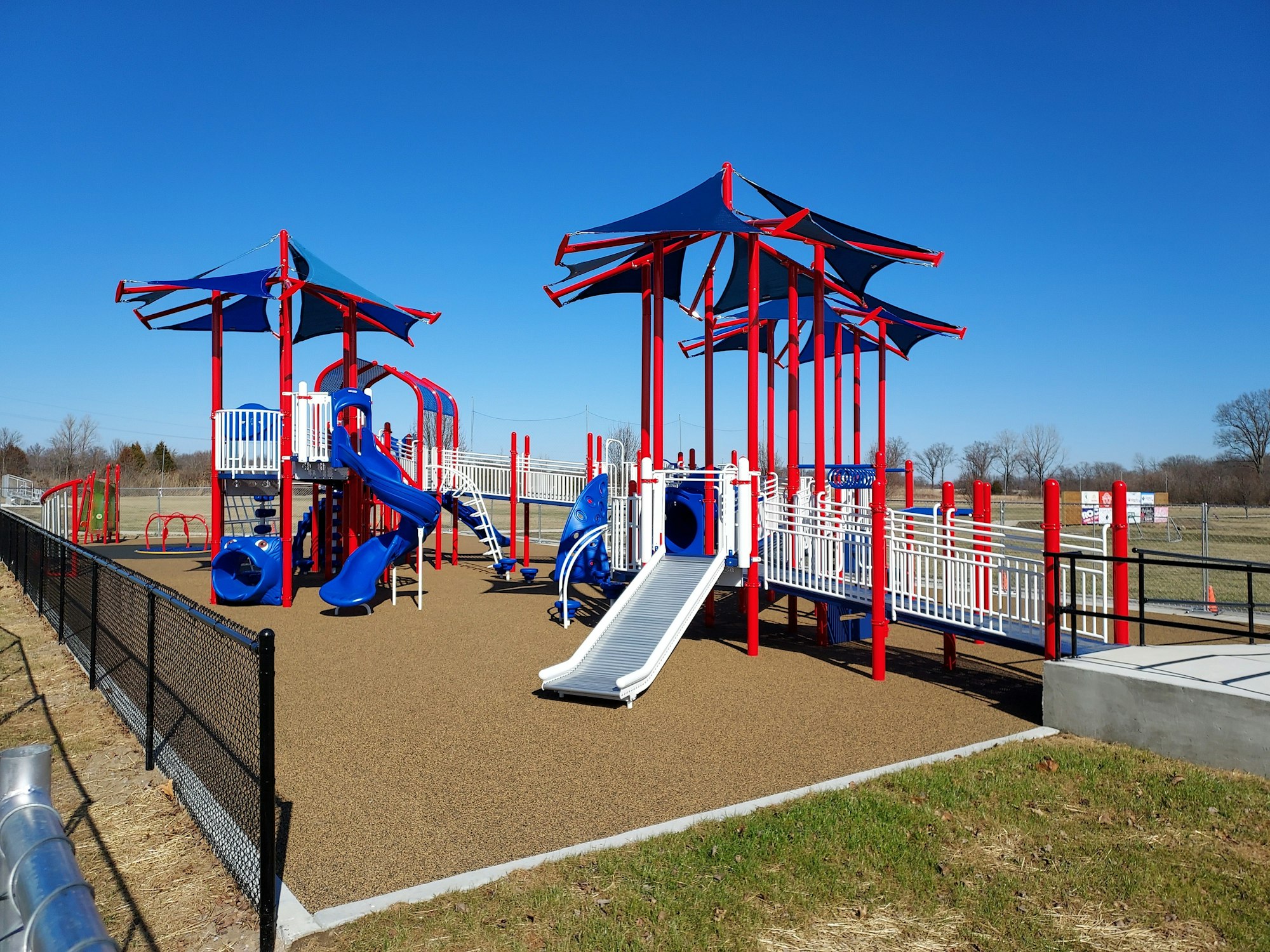 A colorful playground with blue and red structures, slides, and climbing features, surrounded by a safety fence on a sunny day.