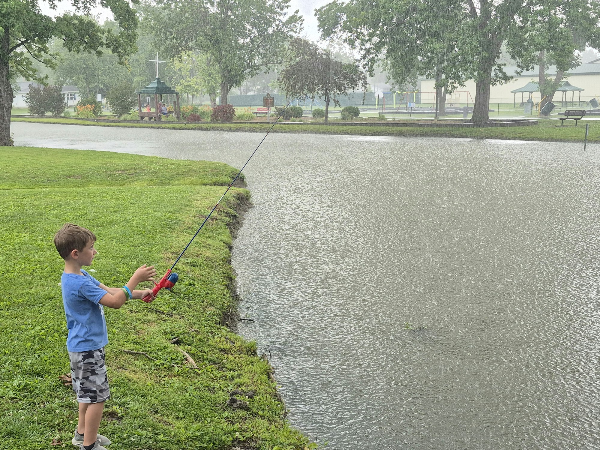A young boy is fishing in the rain at a park, surrounded by greenery and a pond.