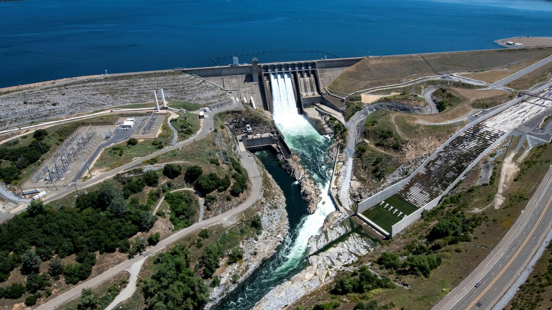 An aerial view of a dam with water flowing through spillways, adjacent roads, power lines, and surrounding landscape.