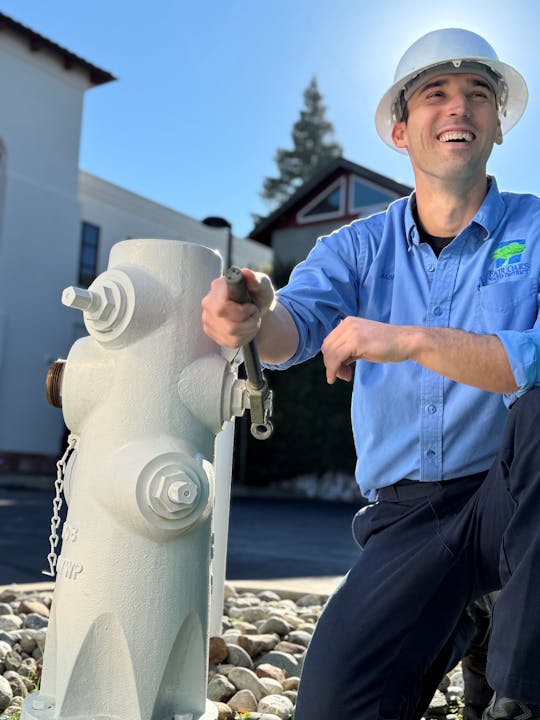 A person in a blue uniform and hard hat is smiling while holding a tool near a fire hydrant outdoors.