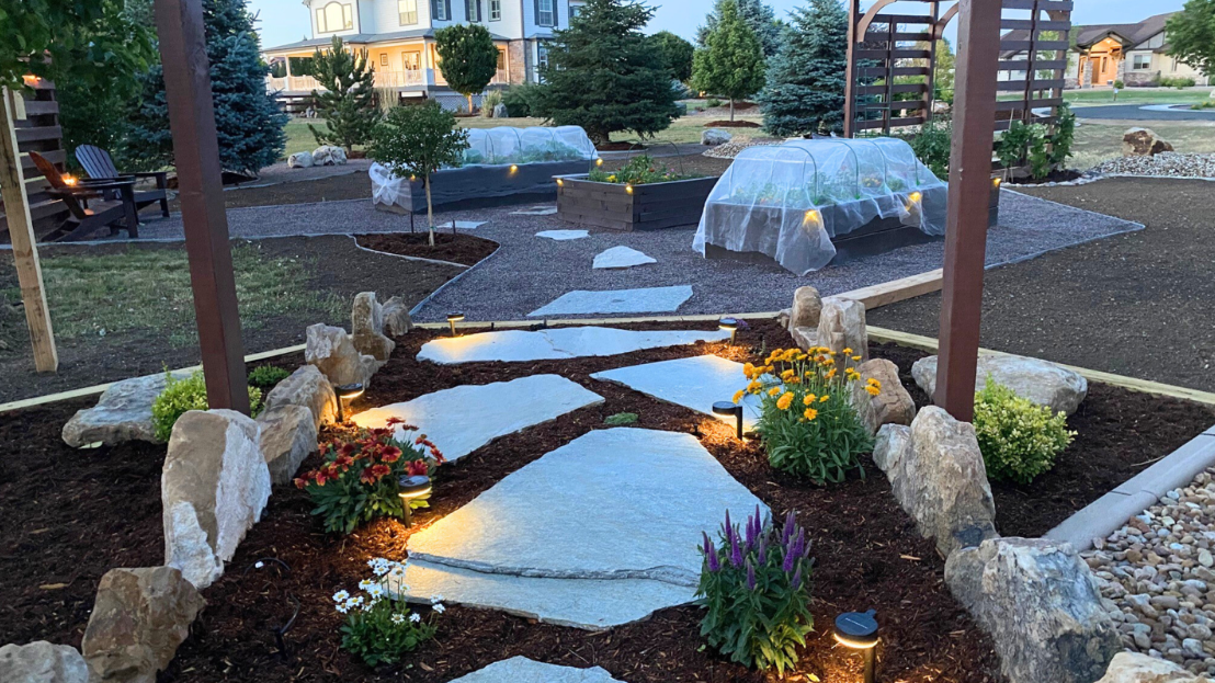 Garden with stone path, flowers, landscape lighting, raised beds with covers, and trees, in front of a house.