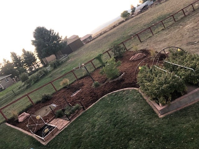 A garden with plants, mulch, and a trellis, bordered by a fence, countryside in the background.