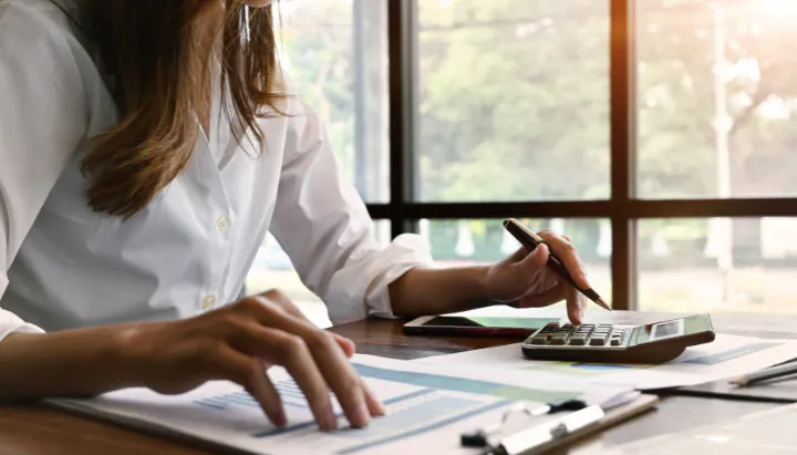 Person using a calculator and reviewing documents at a desk by a large window.