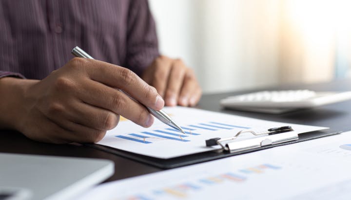 A person analyzing graphs on a clipboard with a pen, alongside a laptop and a blurred keyboard in the background.