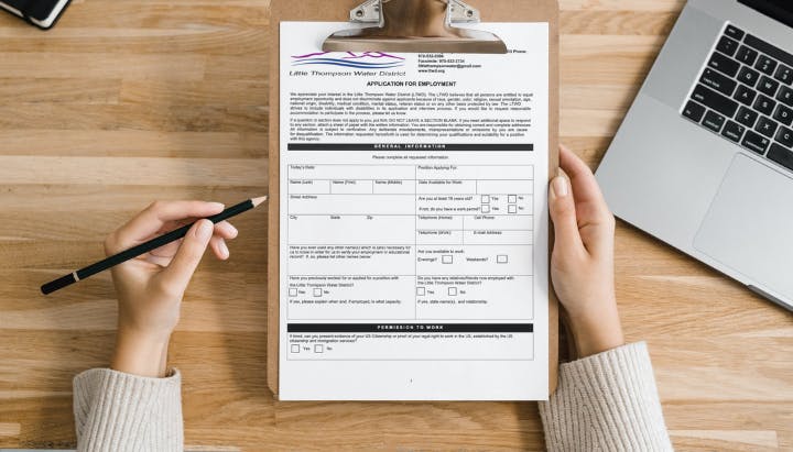 A person holds a clipboard with an employment application form. A pencil is in one hand, and a laptop is nearby on a wooden table.