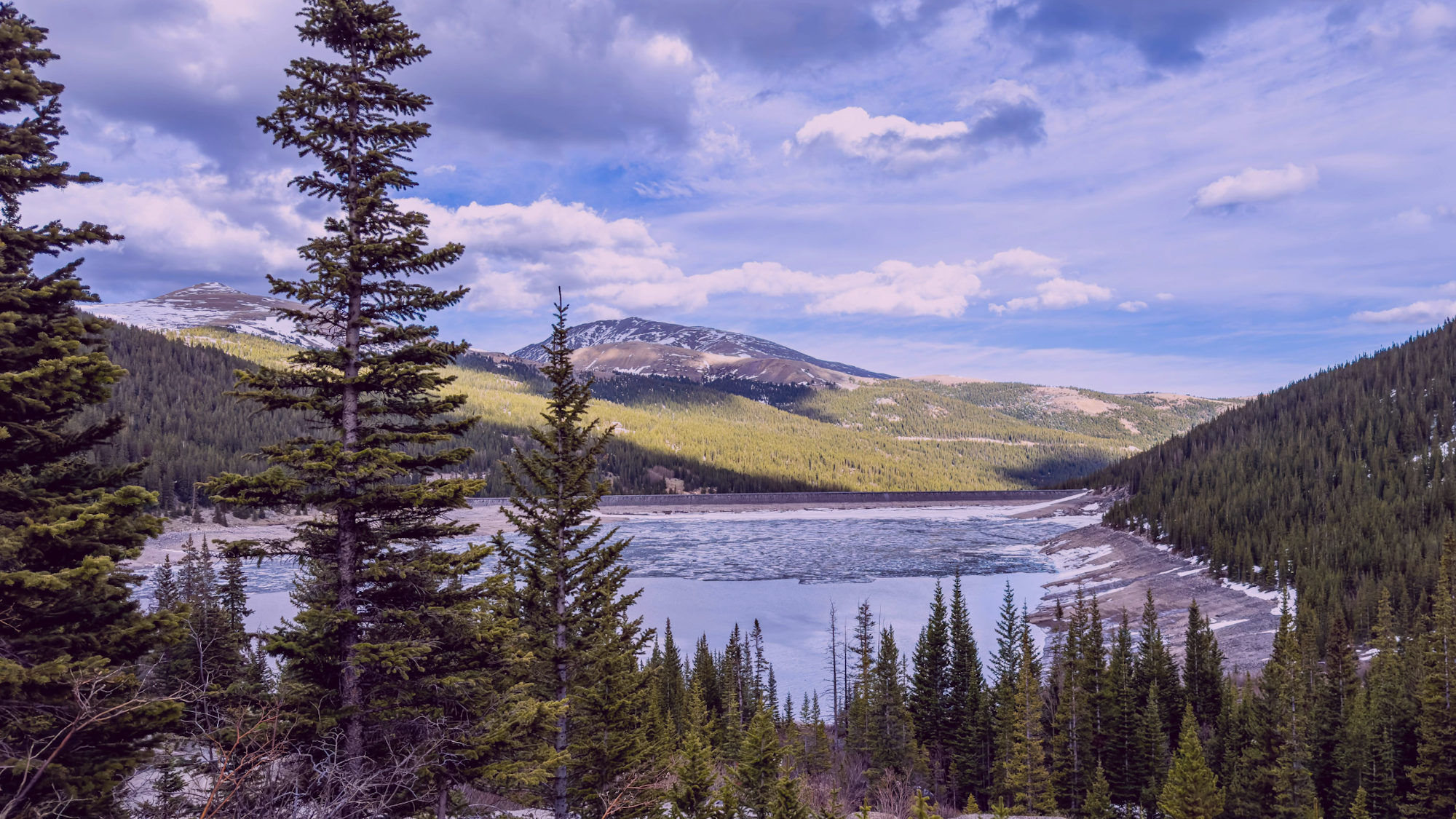 A serene landscape featuring a lake surrounded by evergreen trees, mountains, and a cloudy sky, suggesting tranquility and nature's beauty.