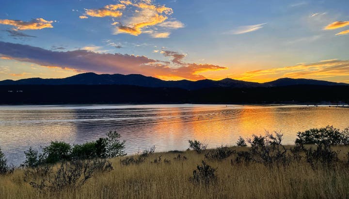 Sunset over a tranquil lake with silhouetted mountains and golden clouds, viewed from a grassy shoreline.