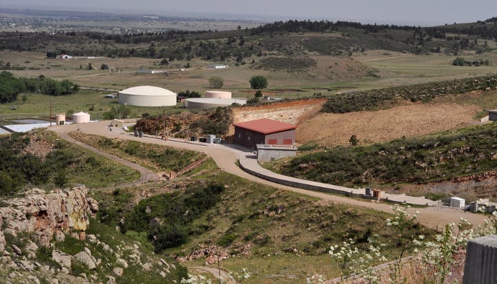 A landscape view with a few buildings, storage tanks, a winding road, and grassy hills in the background.