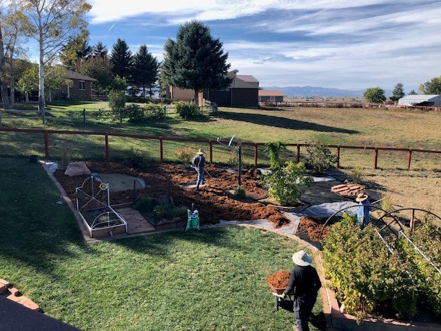 Two people working in a garden with tools and wheelbarrows, surrounded by a fence and trees, open landscape beyond.