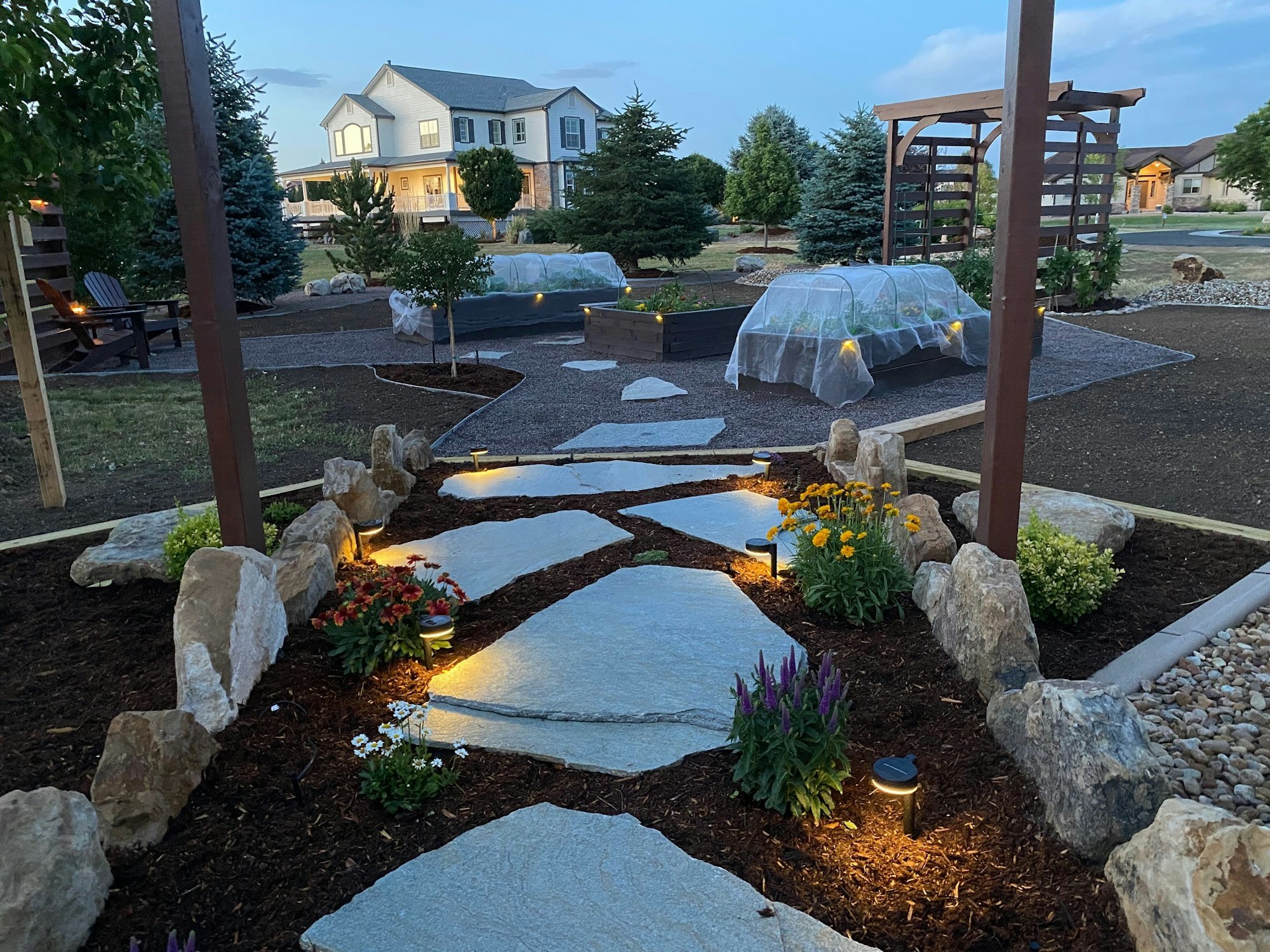 A landscaped garden with stone path, flowers, garden beds, and a house in the background at dusk.