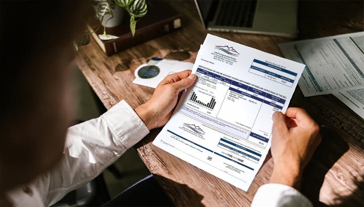 Person holding and examining a utility bill while seated at a desk with a laptop and documents.