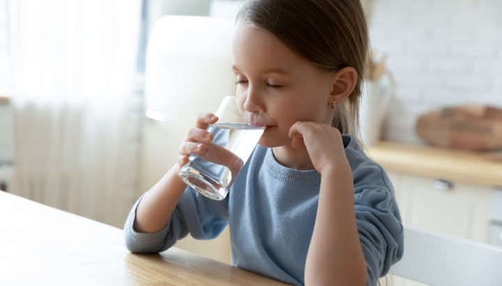 A child in a blue shirt is sitting at a table, drinking water from a glass.