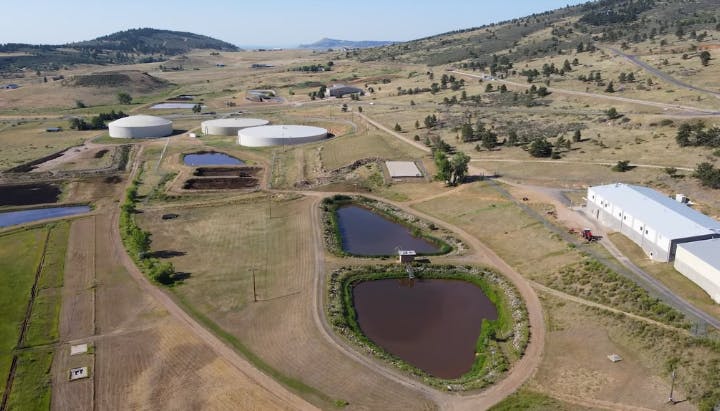 Aerial view of a rural area with large white tanks, rectangular ponds, buildings, and surrounding grassy fields and hills.