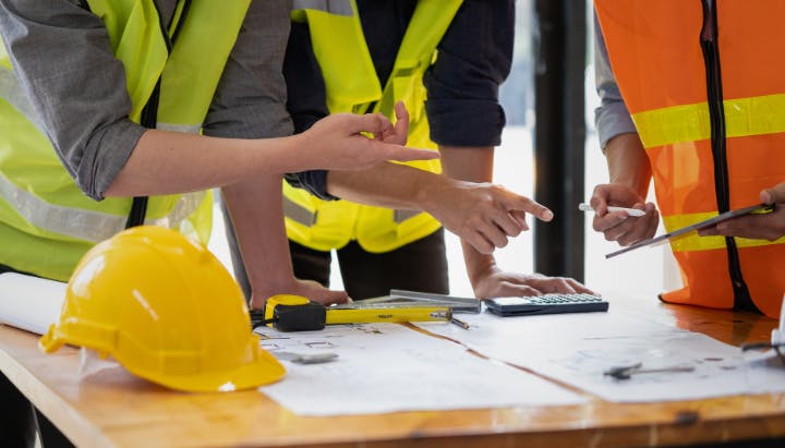 People in safety vests discussing plans at a table with blueprints, a hard hat, and a calculator.