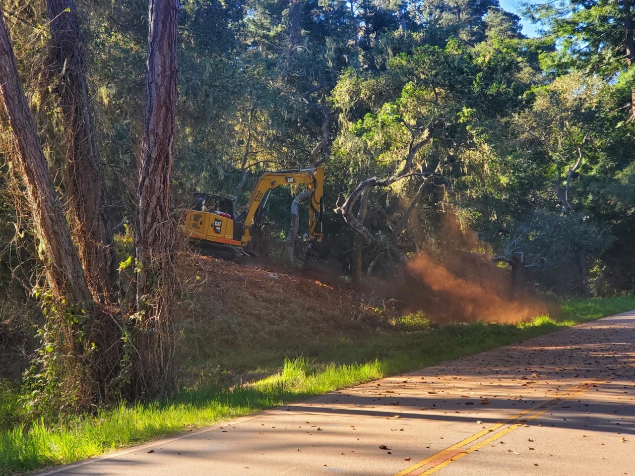 A yellow excavator is working on clearing land near a road, with dust and debris being kicked up from the ground.