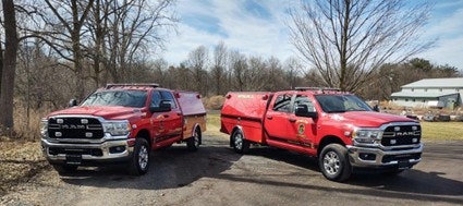 Two red trucks parked outdoors, likely for emergency services, with a background of trees and a building in the distance.
