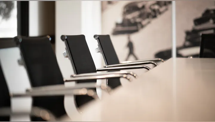 The image shows modern black office chairs lined up next to a sleek table, with a blurred backdrop of a cityscape.