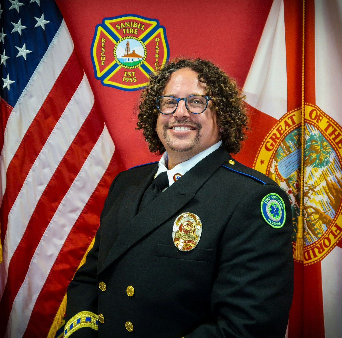 A person in a formal uniform stands in front of U.S. and Florida state flags, with a "Sanibel Fire Rescue District" emblem in the background.