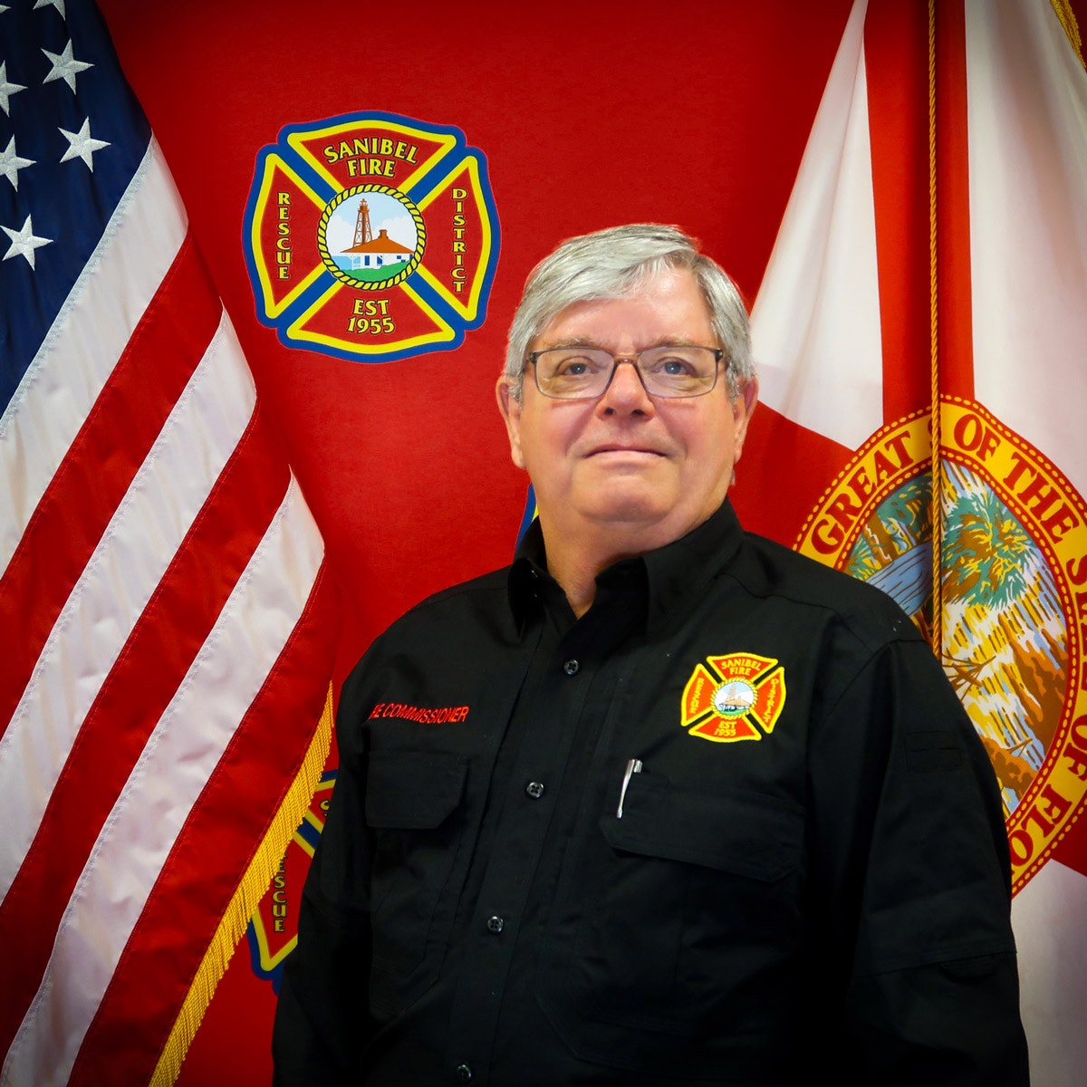 Man in a black uniform stands by American and Florida flags, with a Sanibel Fire Rescue District emblem on the wall.