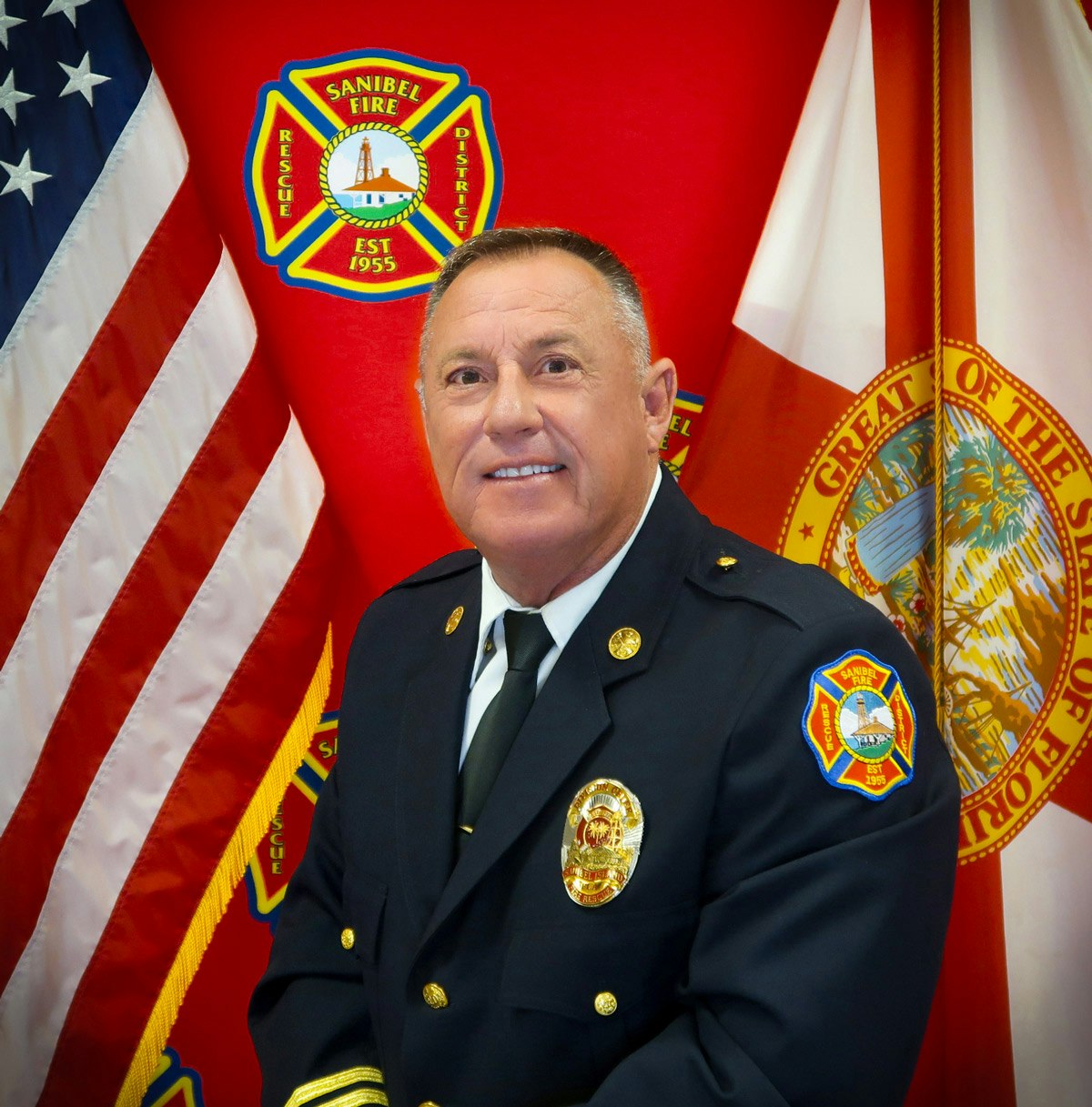 A uniformed officer stands in front of U.S. and Florida flags, with a "Sanibel Fire" emblem visible.