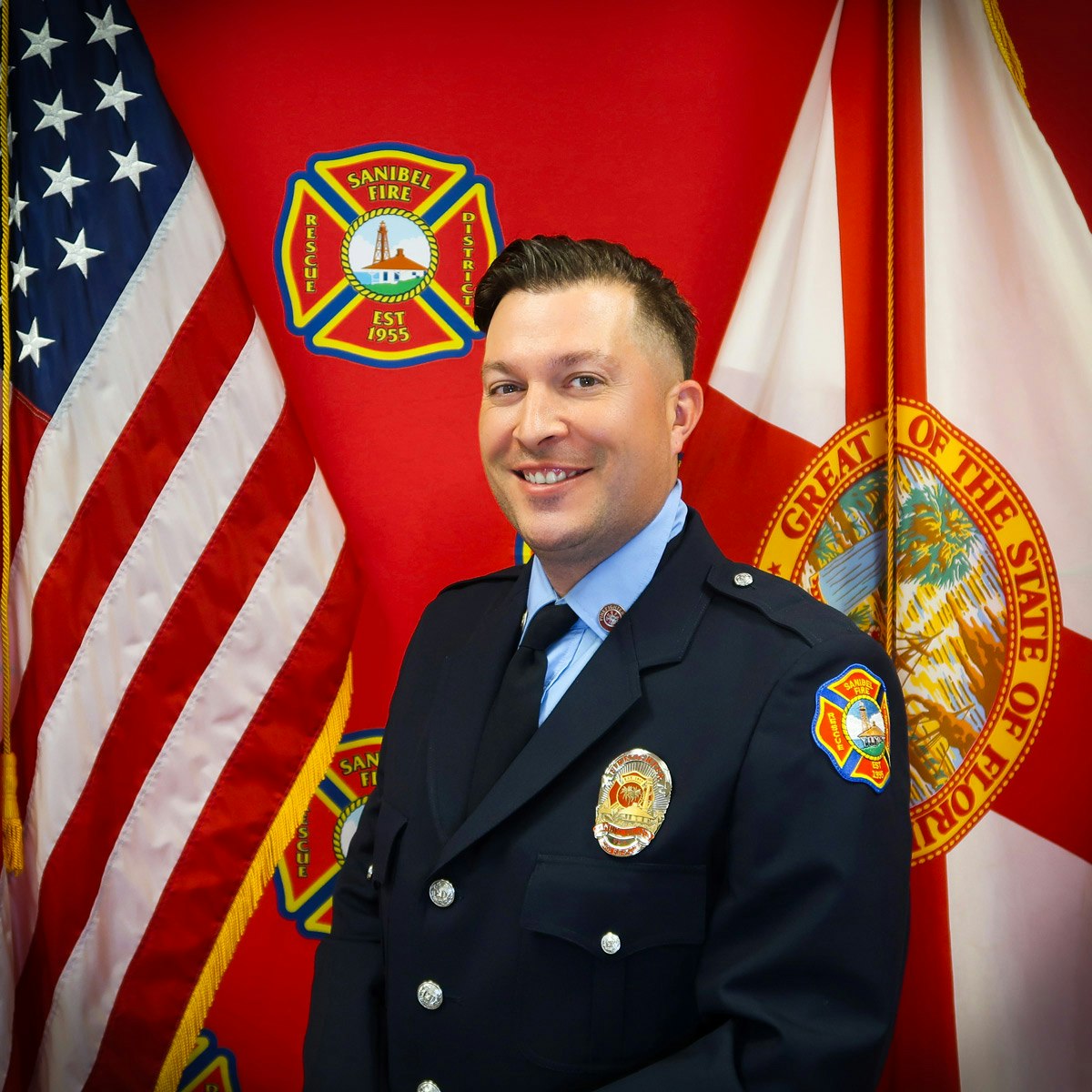 A firefighter in uniform poses in front of US and Florida flags with the Sanibel Fire Rescue District logo.