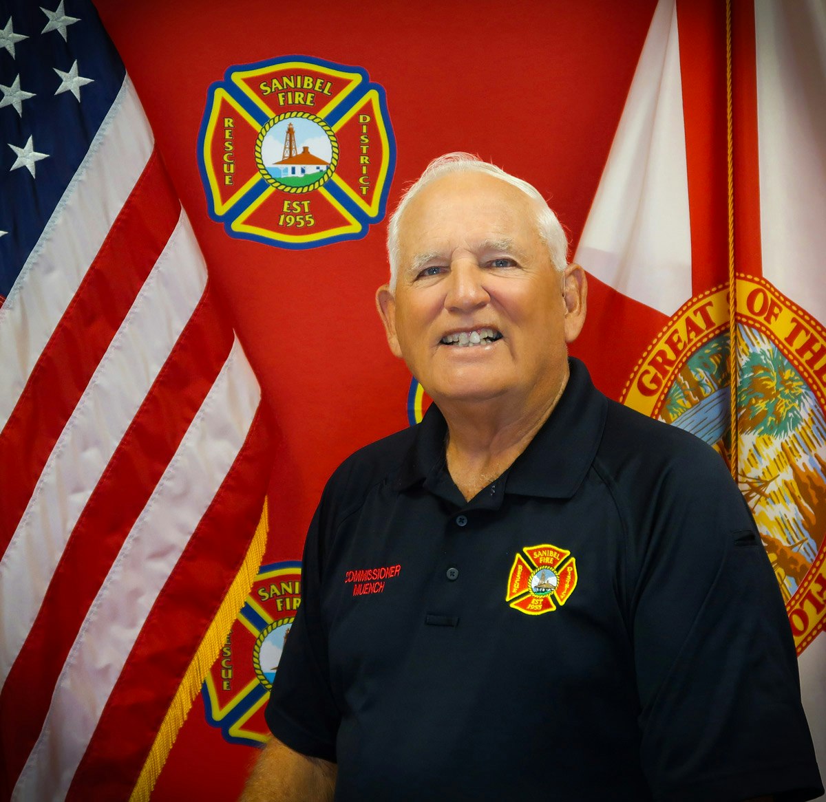 A person in a uniform stands before a Sanibel Fire Rescue District backdrop, flanked by American and state flags.
